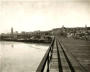 A long wooden boardwalk stretches into the distance towards a hill covered in houses and commercial buildings. A clock tower rises from the horizon. To the left of the boardwalk is shallow water or mud. 